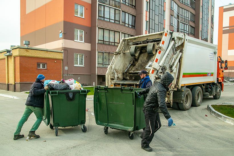 Двое мужчин катят мусорные контейнеры к мусороуборочной машине